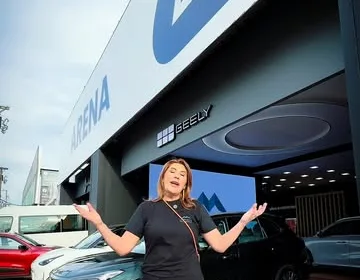 Estande Arena com arco superior azul, letreiro Geely e carros expostos sobre piso de deck de madeira.