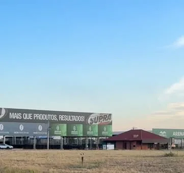 Terreno vazio no parque de exposições, com mato alto e painéis de patrocinadores ao fundo.